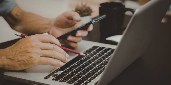 Man holding a pencil in one hand and a mobile phone in the other typing on a laptop at a desk