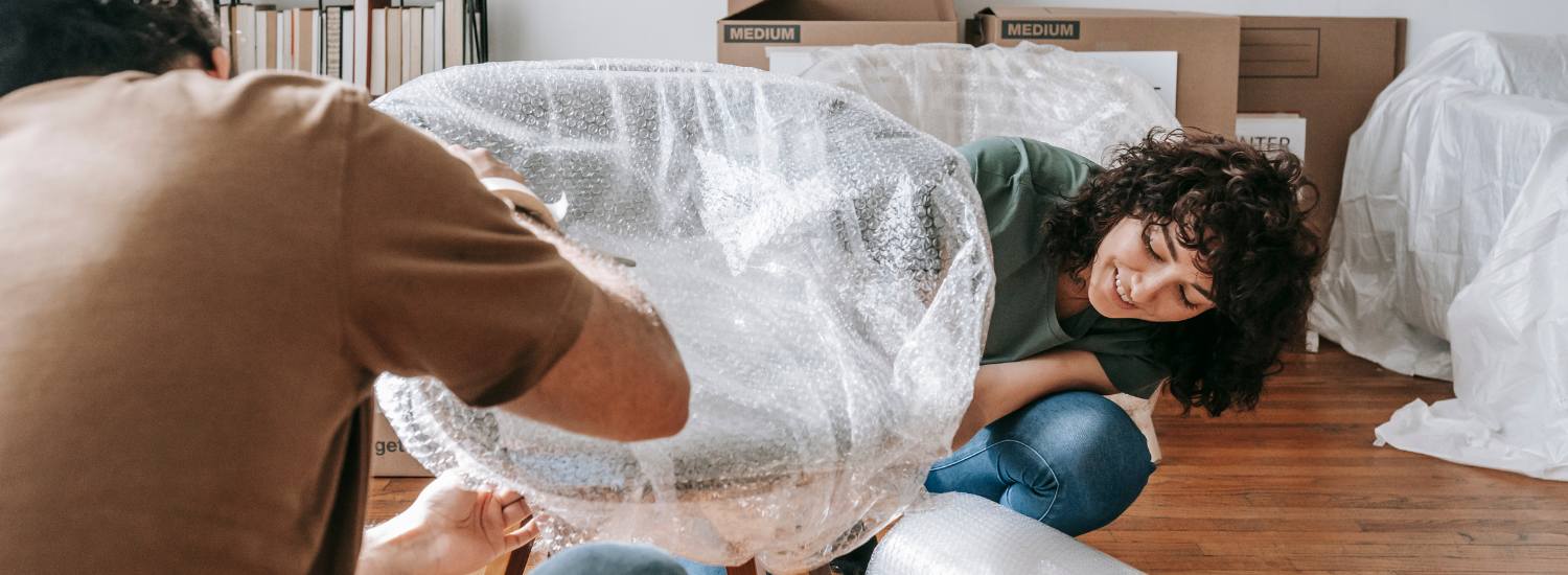 Smiling couple wrapping a small chair in bubble wrap surrounded by moving boxes