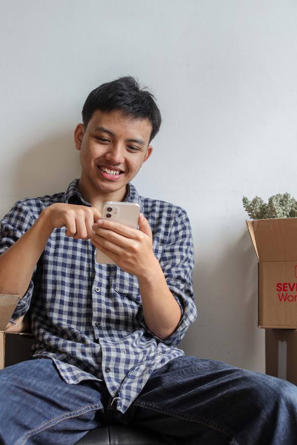 A smiling man in a check shirt next to moving boxes using a mobile phone