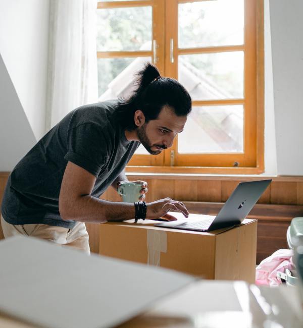 Man checking information on a laptop while packing belongings into cardboard boxes at home.