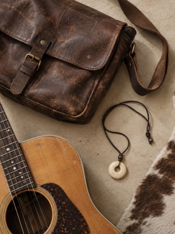 Leather bag, wooden guitar, ivory-detail necklace and hide rug arranged on a neutral surface.